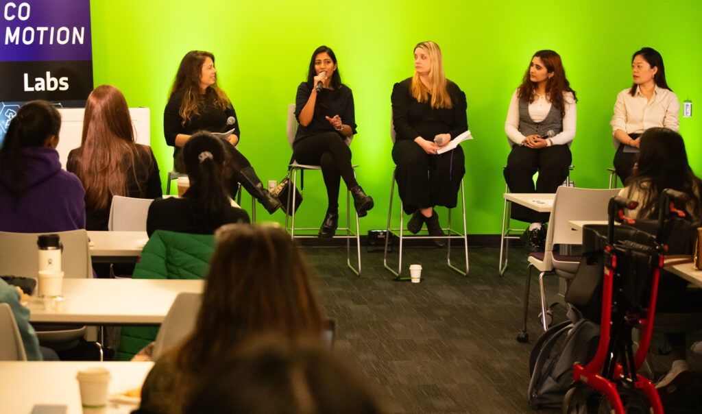A row of women sit on a panel in front of a green screen.