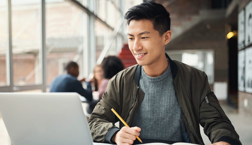 A smiling young man in a modern indoor common space sits at a desk and looks at a laptop computer; he holds a pencil to write in an open notebook beside the computer.