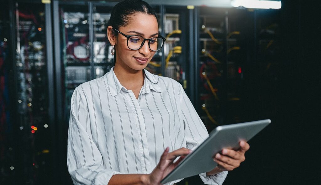 A woman in professional attire stands in a server room, a bank of computers visible in the background; she is using a tablet.