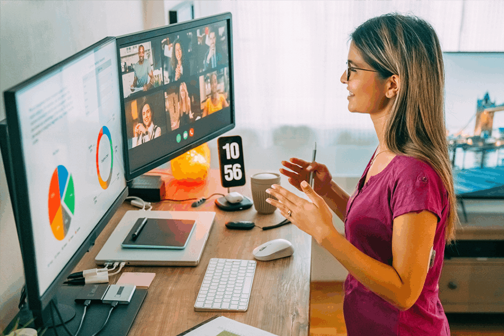 A young woman participates in a video conference call at a desk with multiple monitors displaying charts and meeting participants.