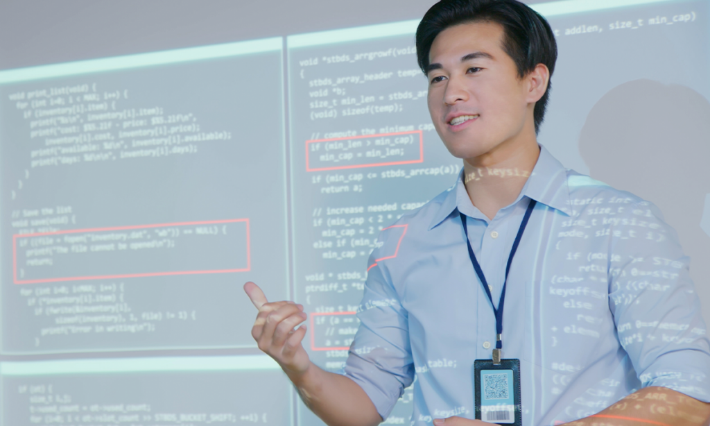 A presenter stands in front of a large screen displaying lines of computer code, with some sections highlighted in red, gesturing as he explains a programming concept.