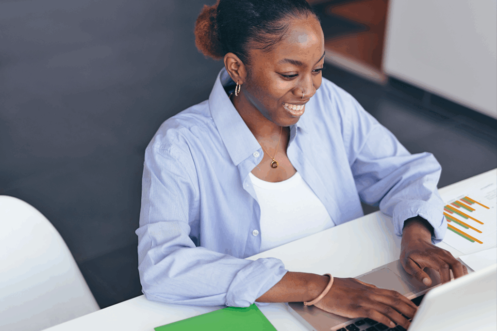 Smiling woman in a light blue shirt, focused and engaged in her work, types on a laptop at a desk with charts and a green folder.