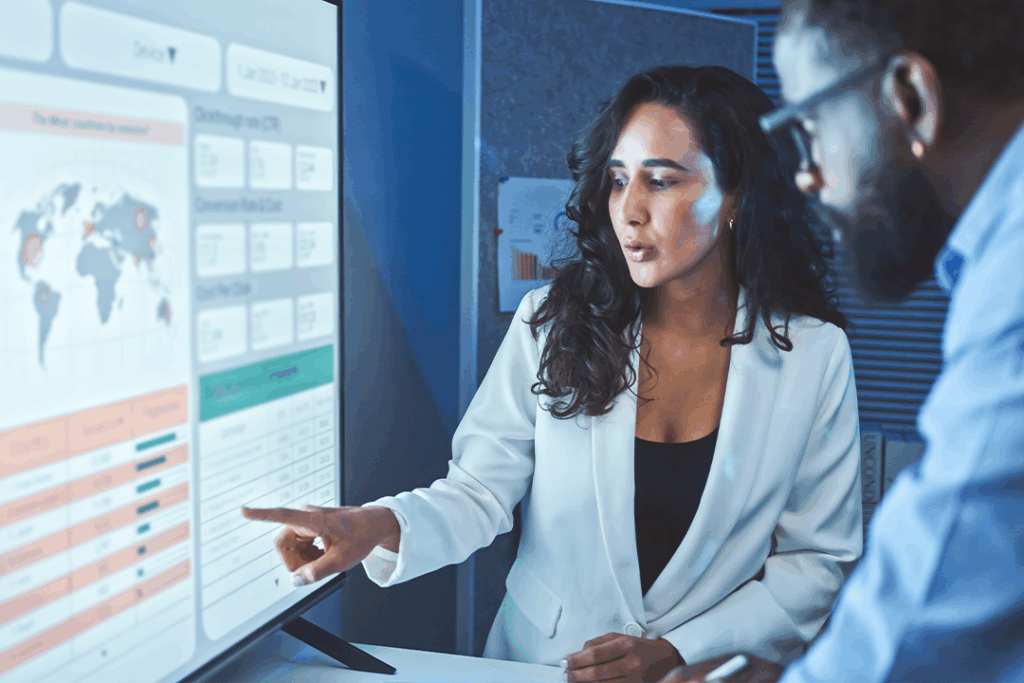 A woman in a white blazer points at a large screen displaying data graphs and a world map, while a man observes attentively beside her.