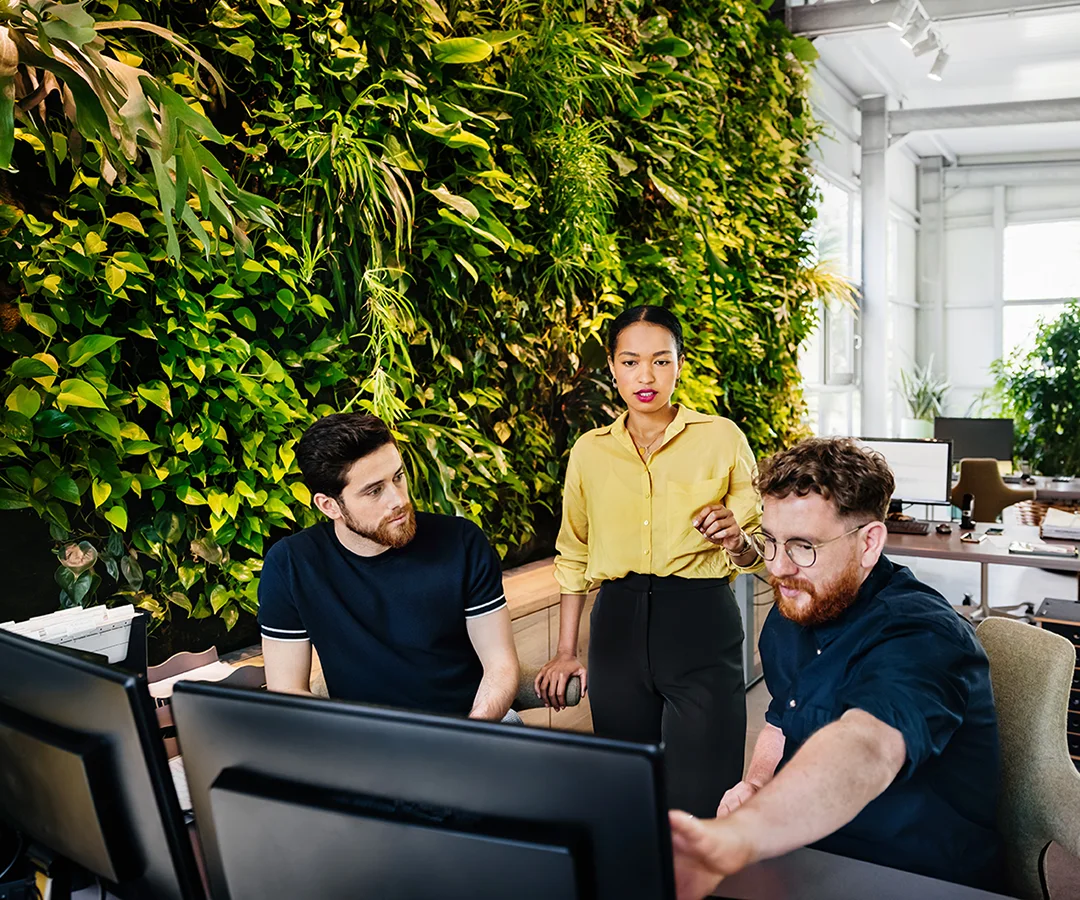 Three professionals smile while looking at a laptop screen in a modern office setting.