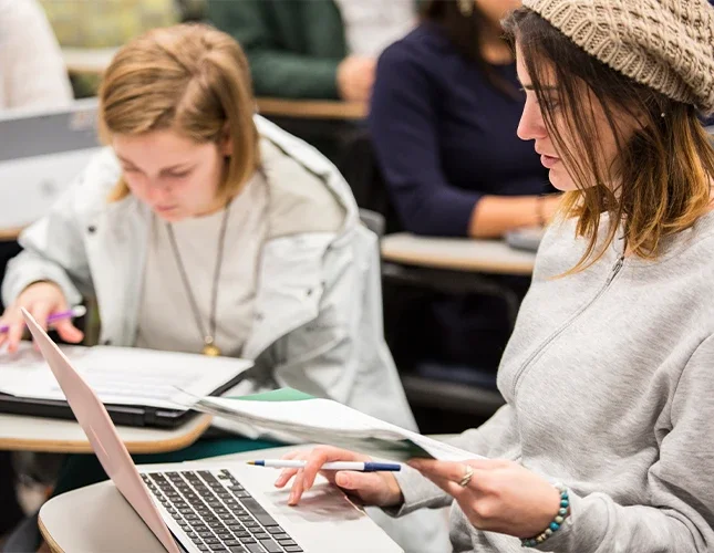 Two MSIM students sit in a lecture hall and learn with a laptop and textbooks.