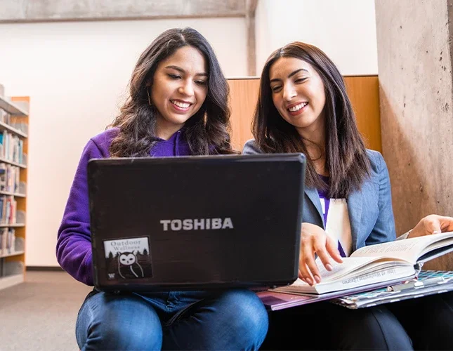 Two MSIM students sit in a library and study with a laptop and textbooks.